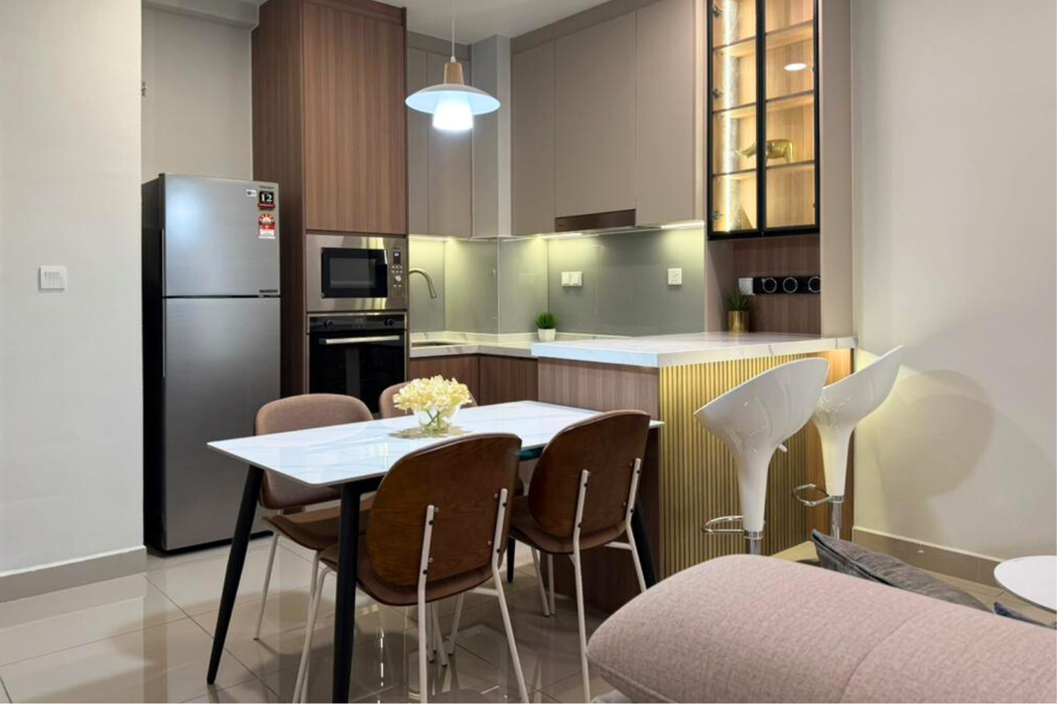 Modern open-concept kitchen with a dining table, surrounded by four chairs, a set of bar stools, and appliances including a refrigerator, microwave, and oven. Stylish decor includes small plants and a hanging light fixture.