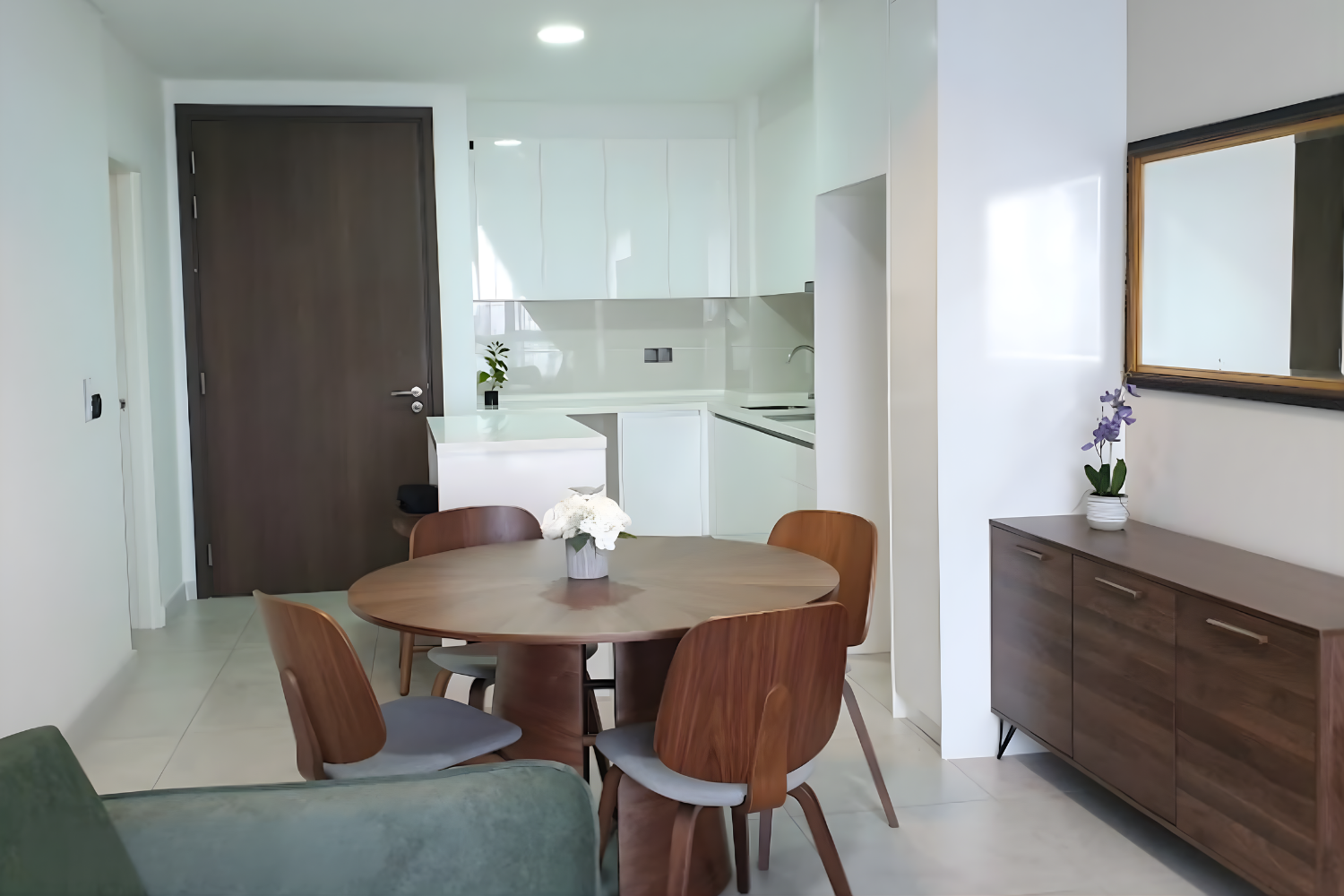 Modern kitchen and dining area with a wooden table, six chairs, white cabinets, a dark door, and a sideboard with a potted purple flower.