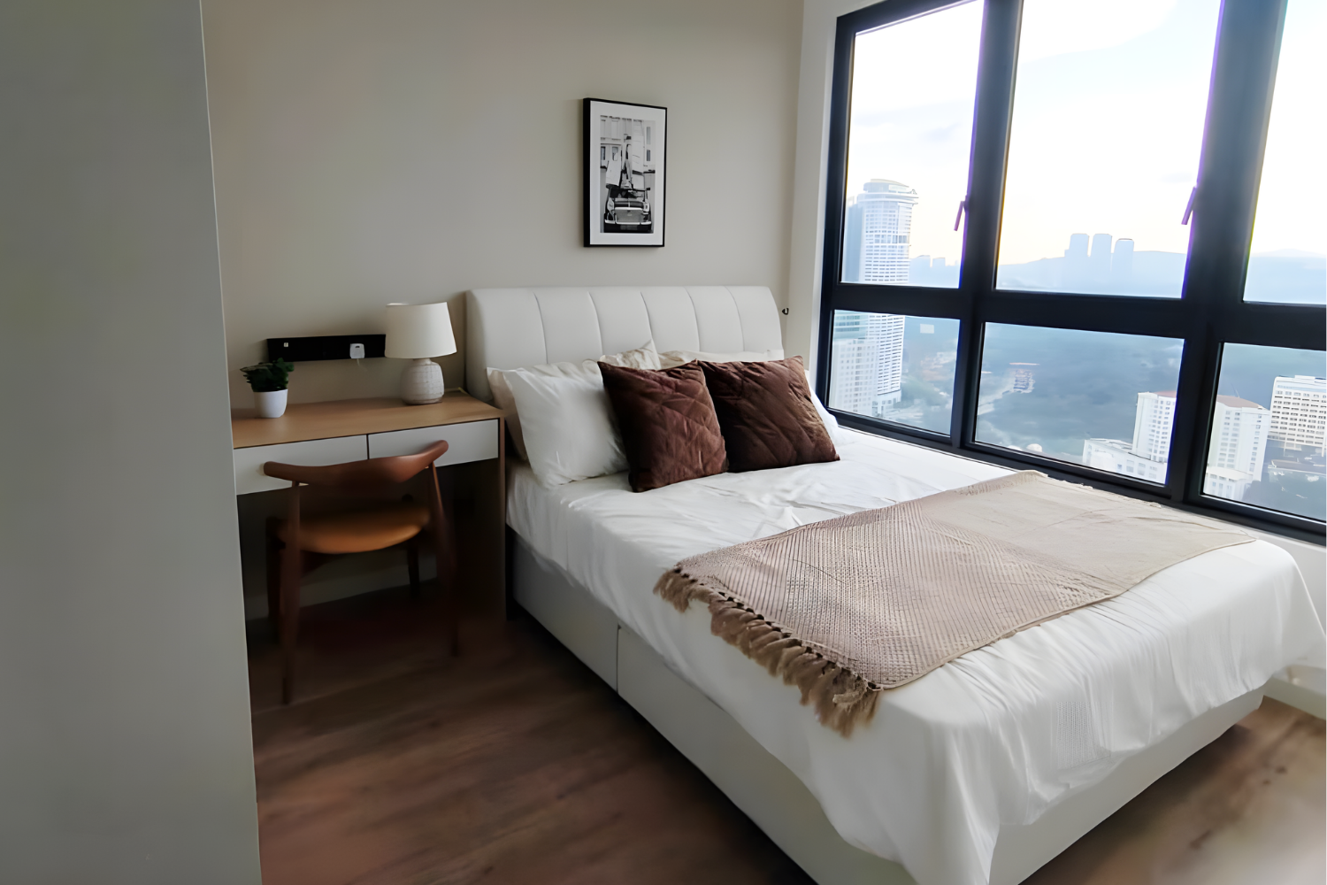A modern bedroom with a large window showing a city skyline, a white bed with brown pillows, a beige throw blanket, a wooden desk with a white drawer, a small lamp, and a framed black-and-white artwork on the wall.