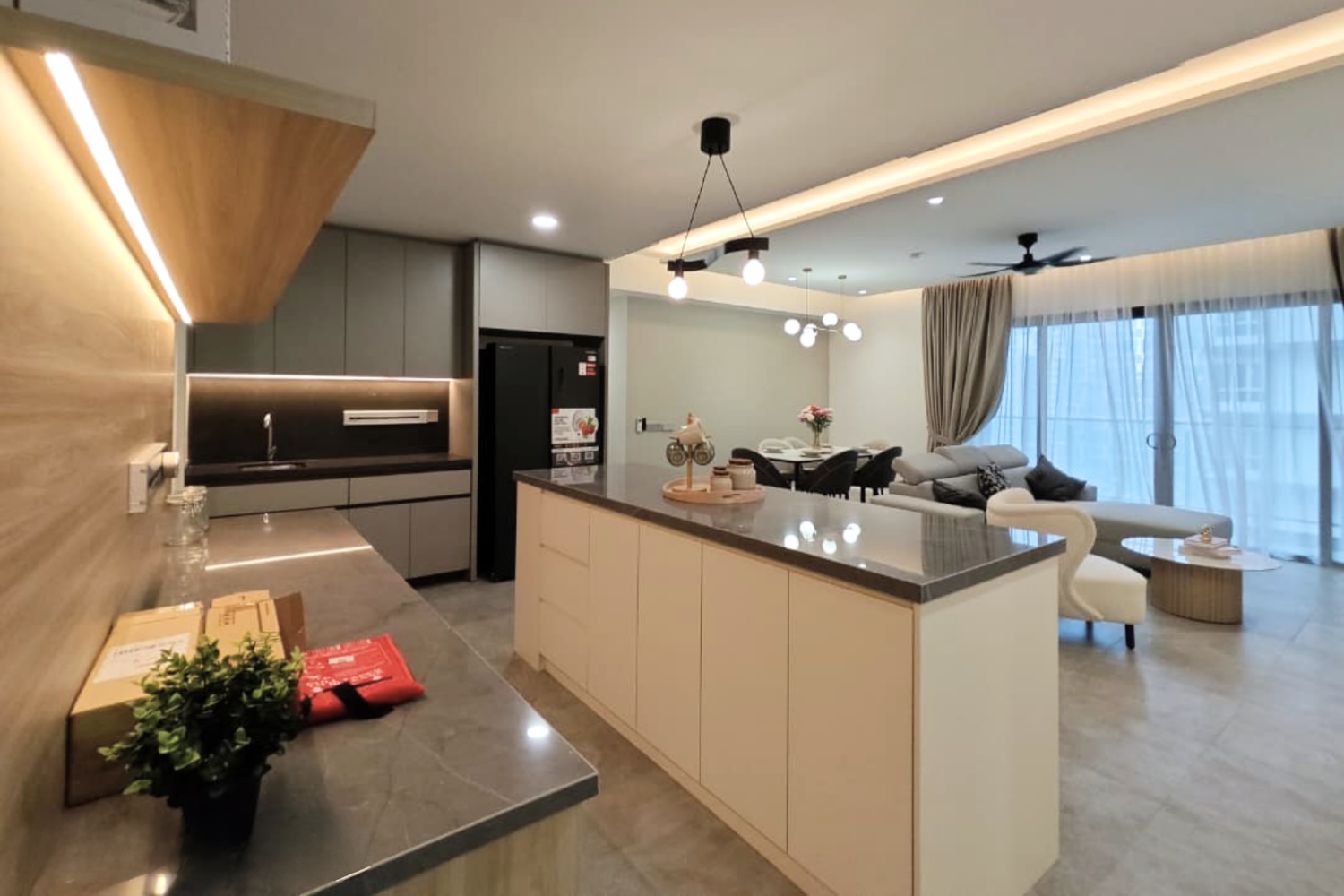 Modern open-plan kitchen and living room with gray cabinets, black refrigerator, island with gray countertop, dining table with black chairs, white sofa, and large glass sliding doors with beige curtains.