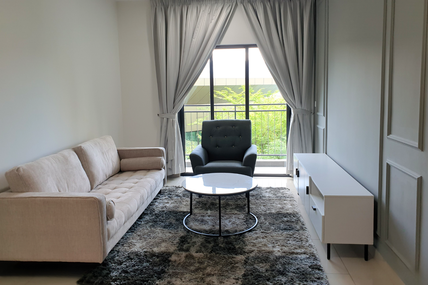 Living room with beige sofa, black armchair, white TV stand, round coffee table, gray shaggy rug, white walls, and large window with gray curtains showing greenery outside.