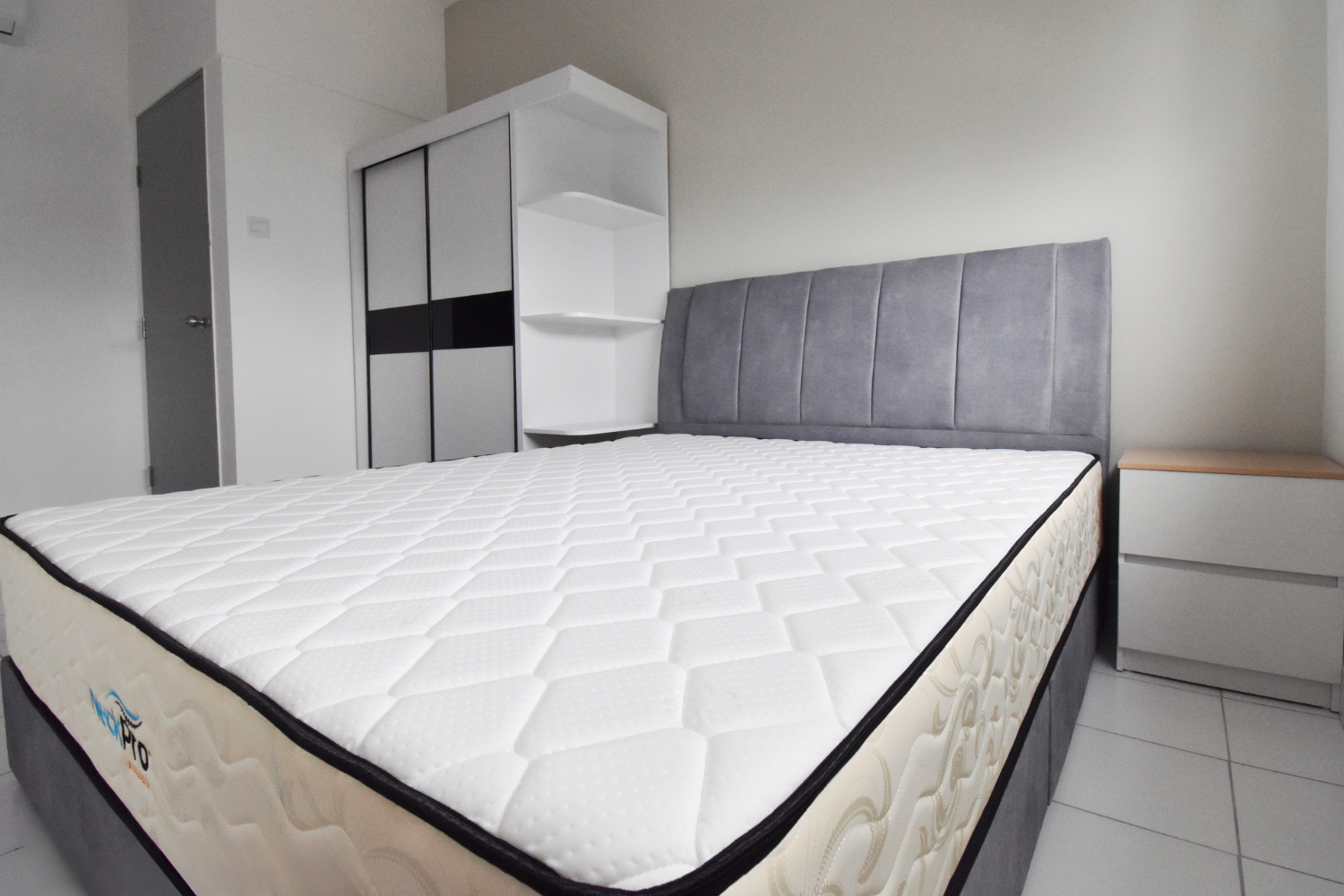 A bedroom with a queen-size bed, gray upholstered headboard, white mattress, white nightstand, closet with sliding doors, and white tiled floor.