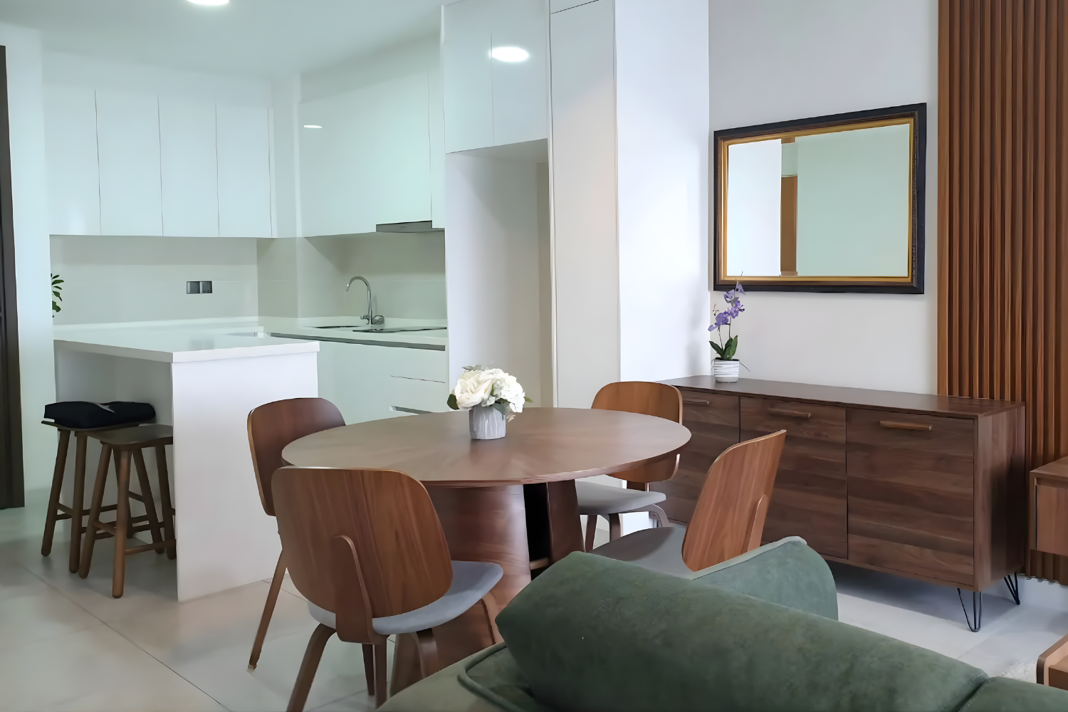 Modern dining area with a round wooden table, six matching chairs, a sideboard with a mirror above, a vase of purple orchids, and a white kitchen with a countertop and sink in the background.