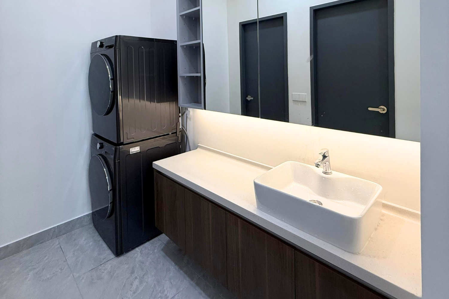 Laundry room with stacked black washer and dryer, white countertop with a large white sink, dark cabinet below, and open shelving above, with three dark doors in the background.