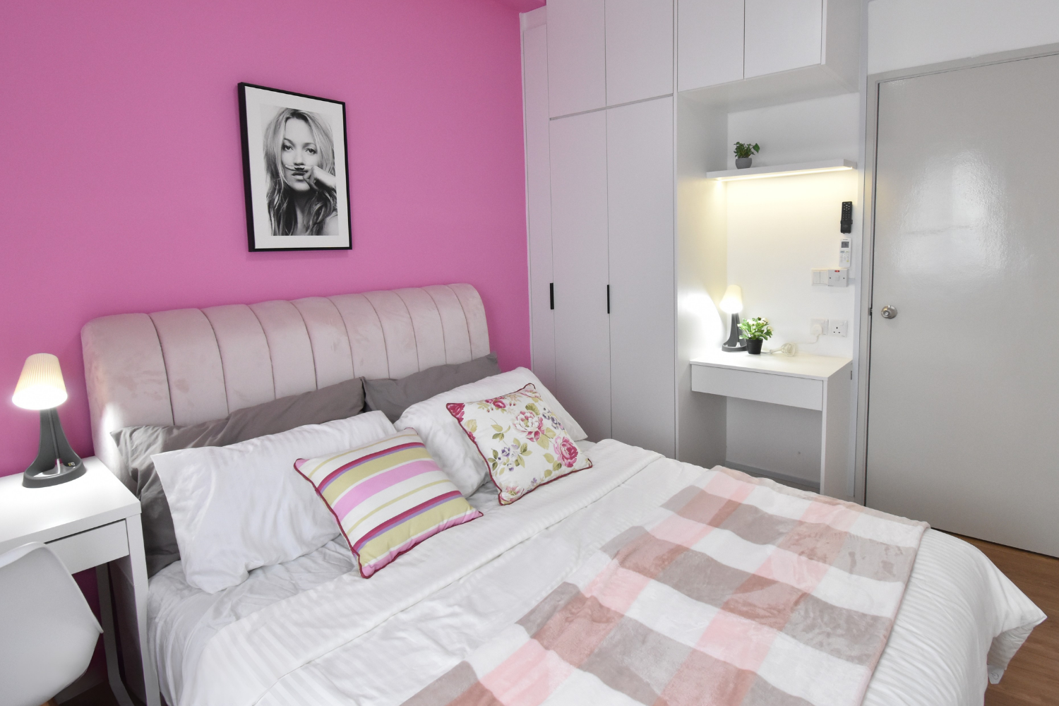 A bedroom featuring a pink accent wall with a black-and-white portrait of a woman, a bed with white bedding, gray and floral pillows, a pink and beige checkered blanket, white bedside tables with lamps, a built-in wardrobe, and a small white desk wit