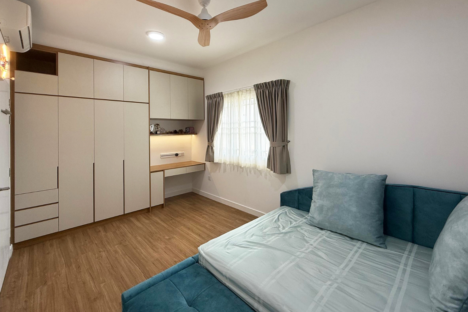 A bedroom with a large built-in white wardrobe, a small desk with shelves, a window with curtains, a ceiling fan, and a blue sofa bed with pillows.