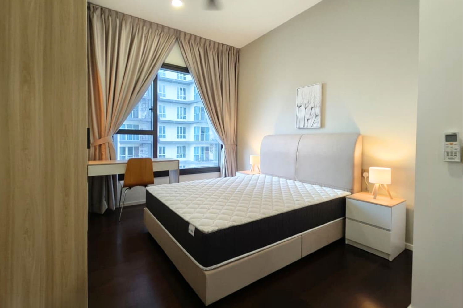 Modern bedroom with a large window, beige bed with headboard, two nightstands with lamps, a desk with a chair, and light-colored curtains.
