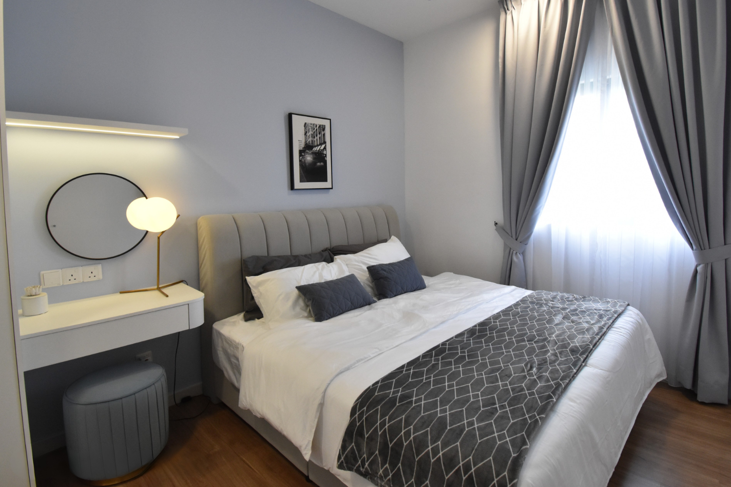 A neatly made bed with white sheets, dark gray pillows, and a patterned gray bed runner in a modern bedroom with light blue walls, a black and white photograph on the wall, and large windows with gray curtains.