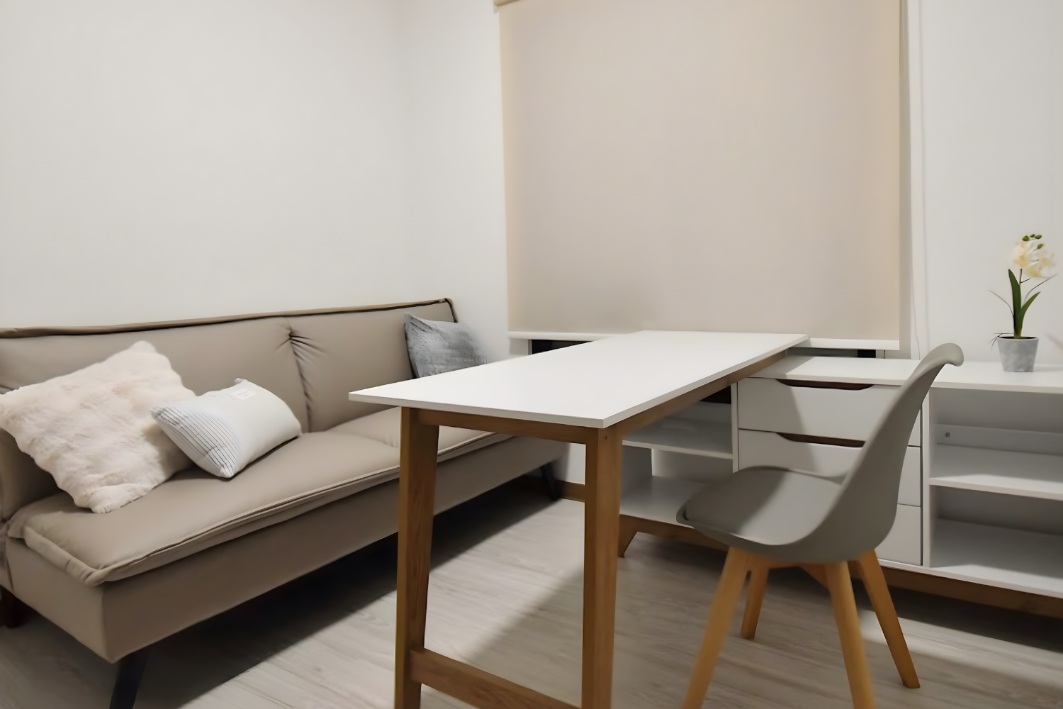 A modern living room with a beige sofa, gray pillows, a white table, gray chair, and a white sideboard with a potted orchid plant.