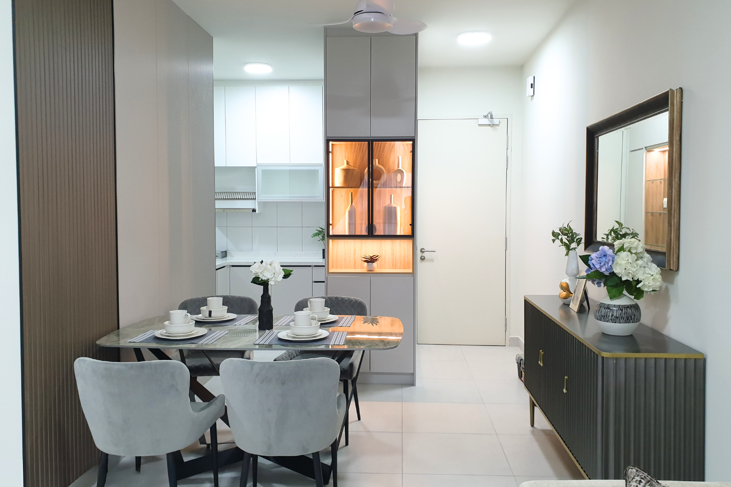 Modern dining area with a marble table set with white dishes, surrounded by four upholstered chairs, next to a gray and gold sideboard with a flower vase, with a mirror and decorative items on top. The background includes a kitchen with white cabinet