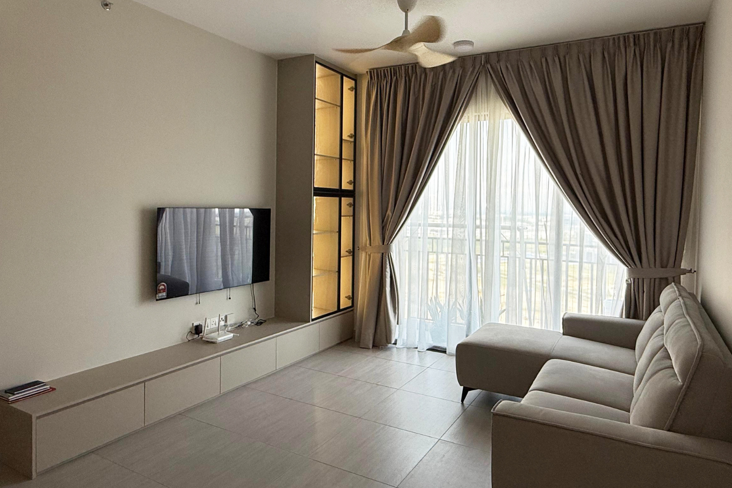 Living room with a beige sectional sofa, a wall-mounted flat-screen TV, a built-in cabinet with glass display shelves, large windows with layered curtains, tiled flooring, and a ceiling fan.