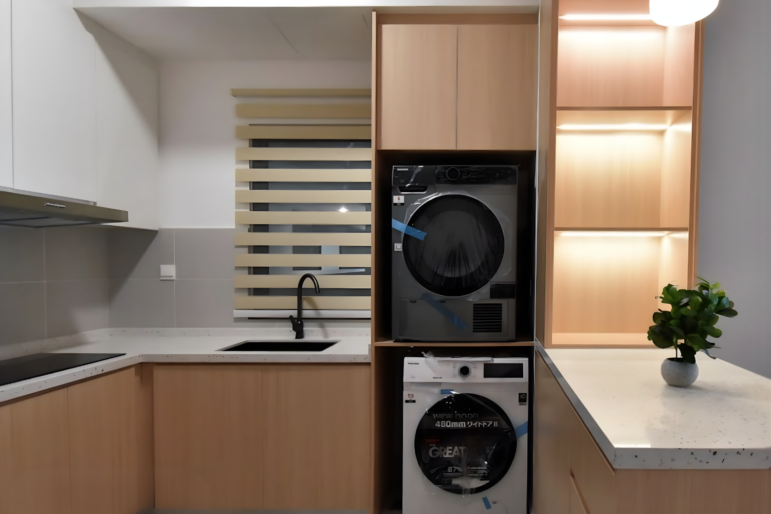 Modern kitchen with light wood cabinets, white countertops, gray backsplash, black faucet, and a small potted green plant on the right. Laundry appliances are integrated into the cabinetry, including a front-loading dryer and washing machine, with a 