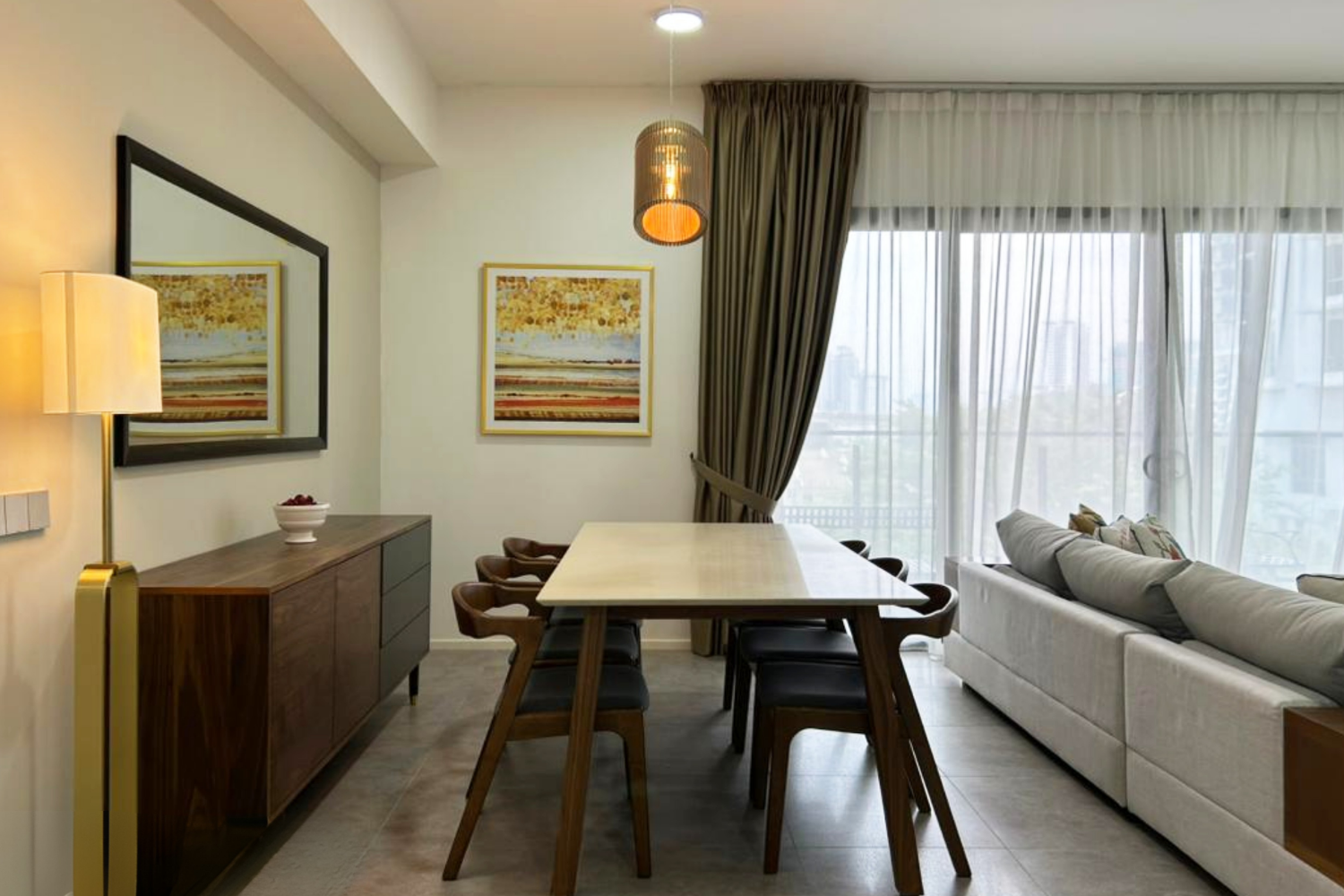 Modern living room with dining area, featuring a wooden dining table, six chairs, a beige sofa, large windows with sheer curtains and dark drapes, artwork on the walls, and a pendant light.