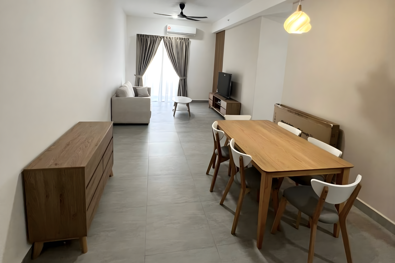 Living room with a sofa, coffee table, TV on a wooden stand, dining table with six chairs, wooden sideboard, and a large window with curtains.