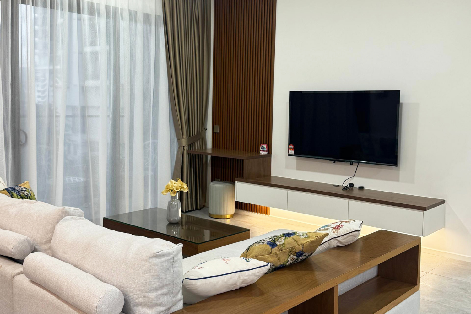 Modern living room with beige sofa, decorative pillows, glass-topped coffee table with vase of yellow flowers, wall-mounted TV, wooden panel accent wall with beige curtains over large window.