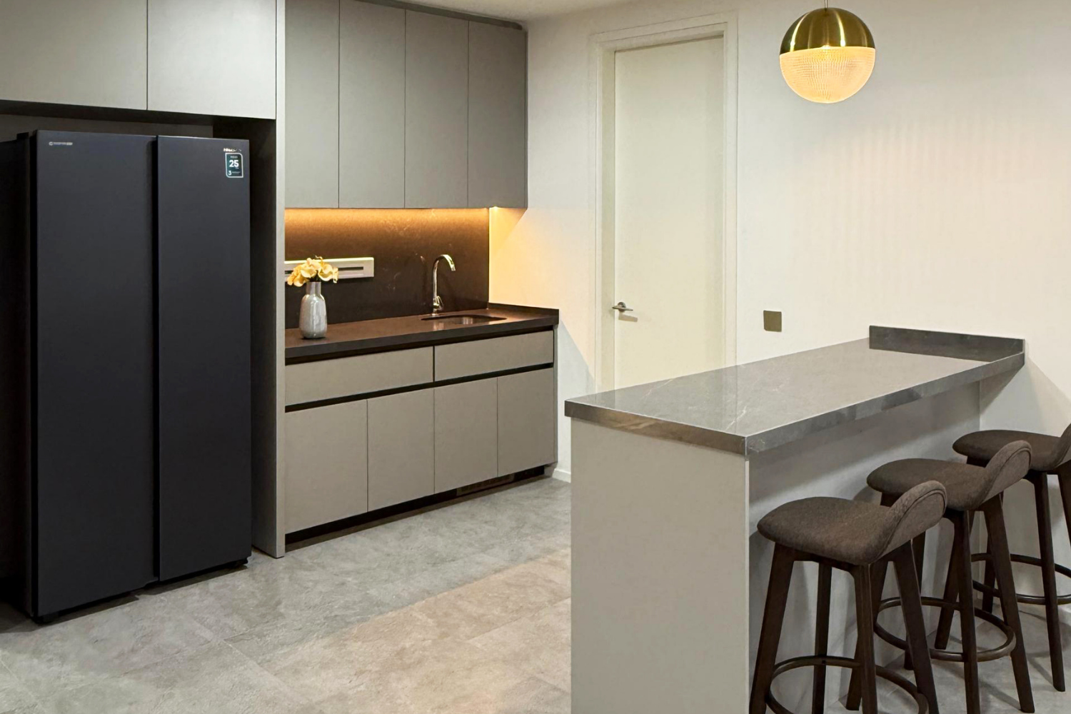 Modern kitchen with gray cabinets, black refrigerator, marble countertop breakfast bar, bar stools, and a gold hanging pendant light.