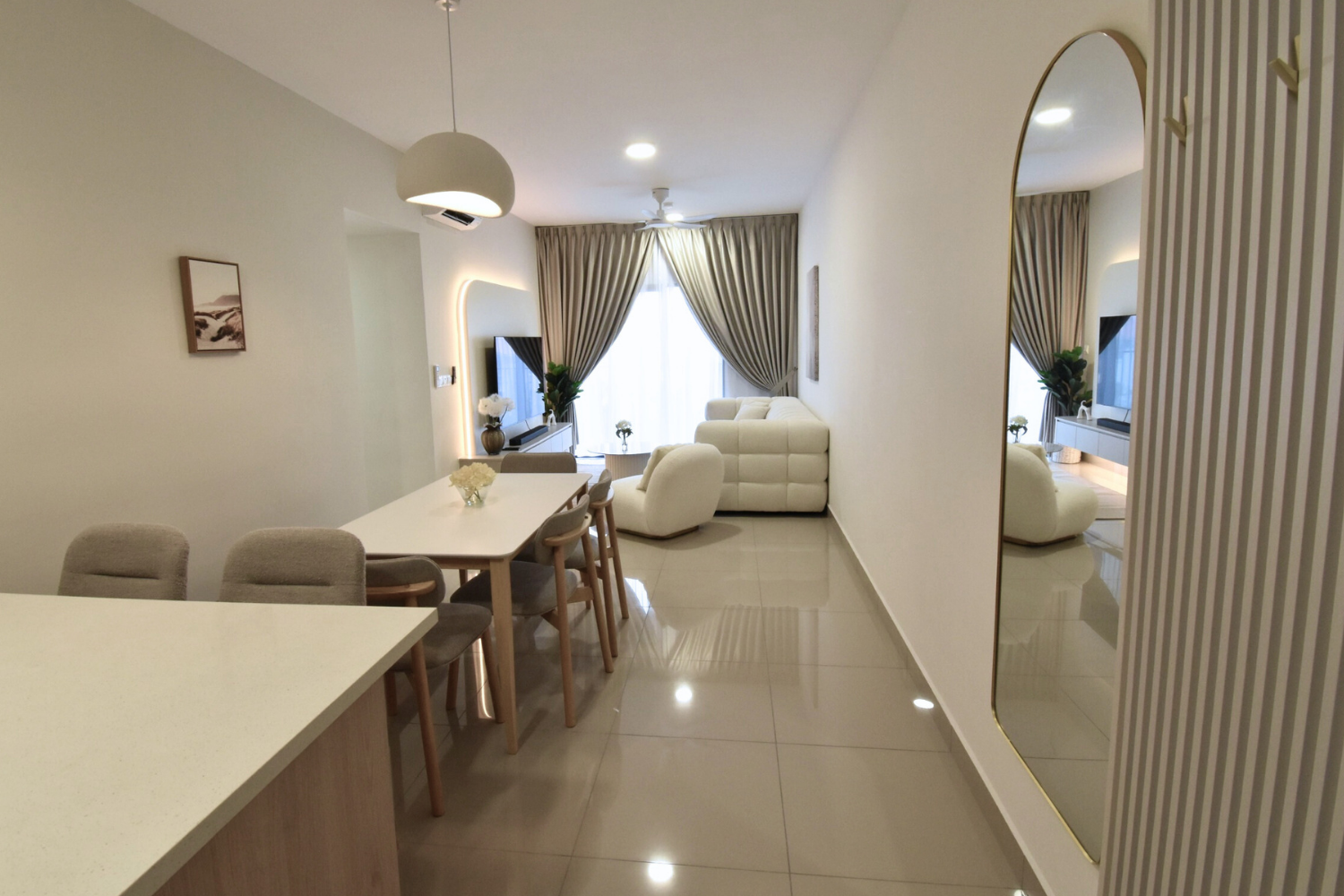 Living room and dining area with modern furniture, beige chairs, white sofa, velvet curtains, wall-mounted TV, large mirror, and decorative plants.