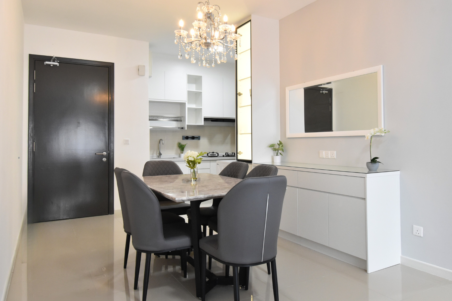Modern dining area with a marble table, six grey chairs, a white sideboard with a mirror, and decorative plants, adjacent to a kitchen with white cabinets and a chandelier.