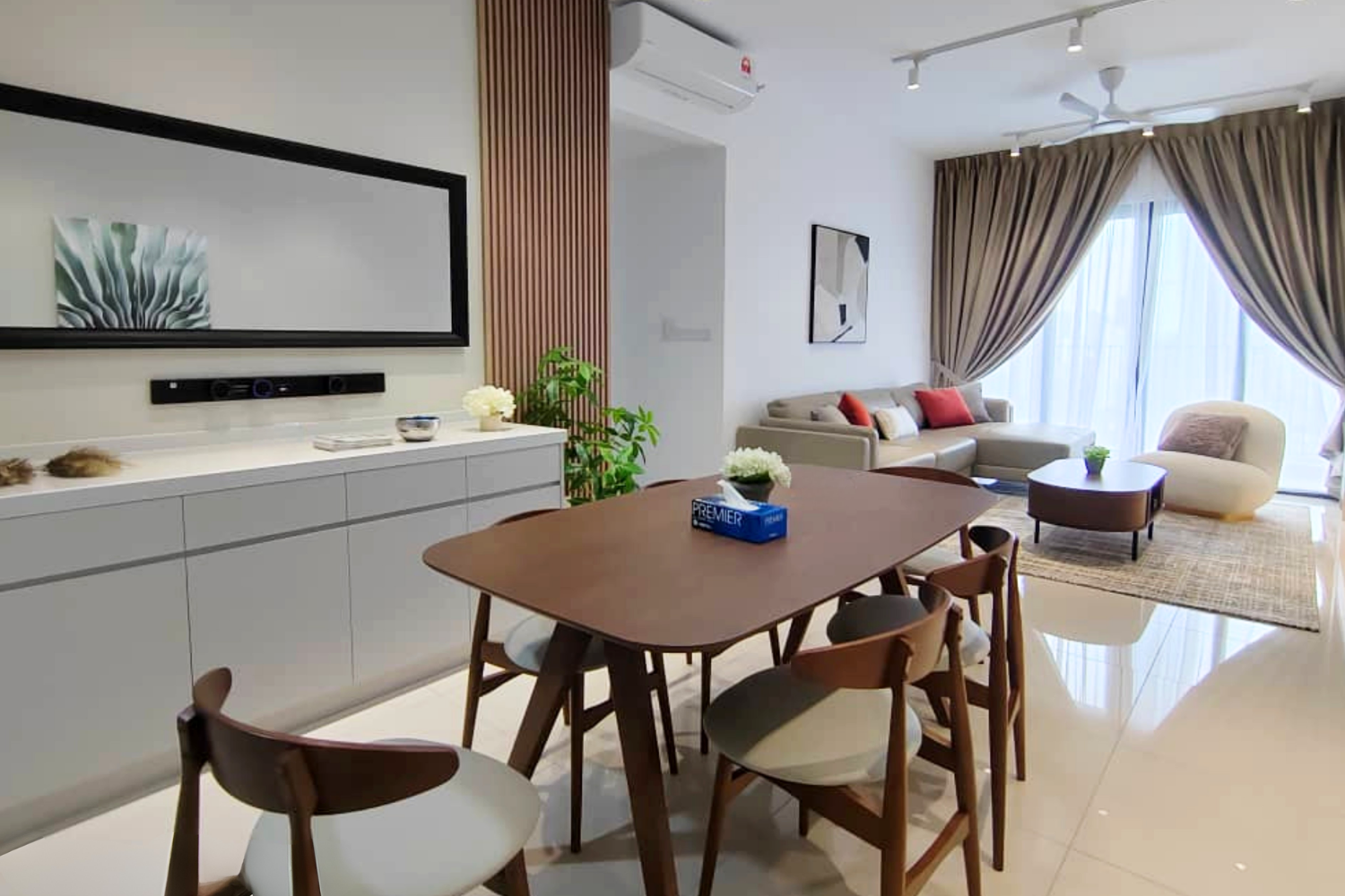 Modern living room and dining area with beige sofas, wooden dining table, chairs, large window with curtains, wall art, and indoor plants.