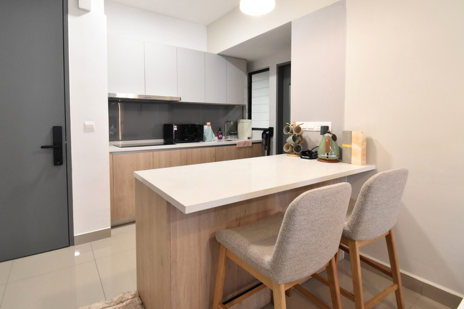 Modern kitchen with white and gray cabinets, a white countertop center island with two beige upholstered chairs, various small appliances, mugs, and supplies on the countertop.