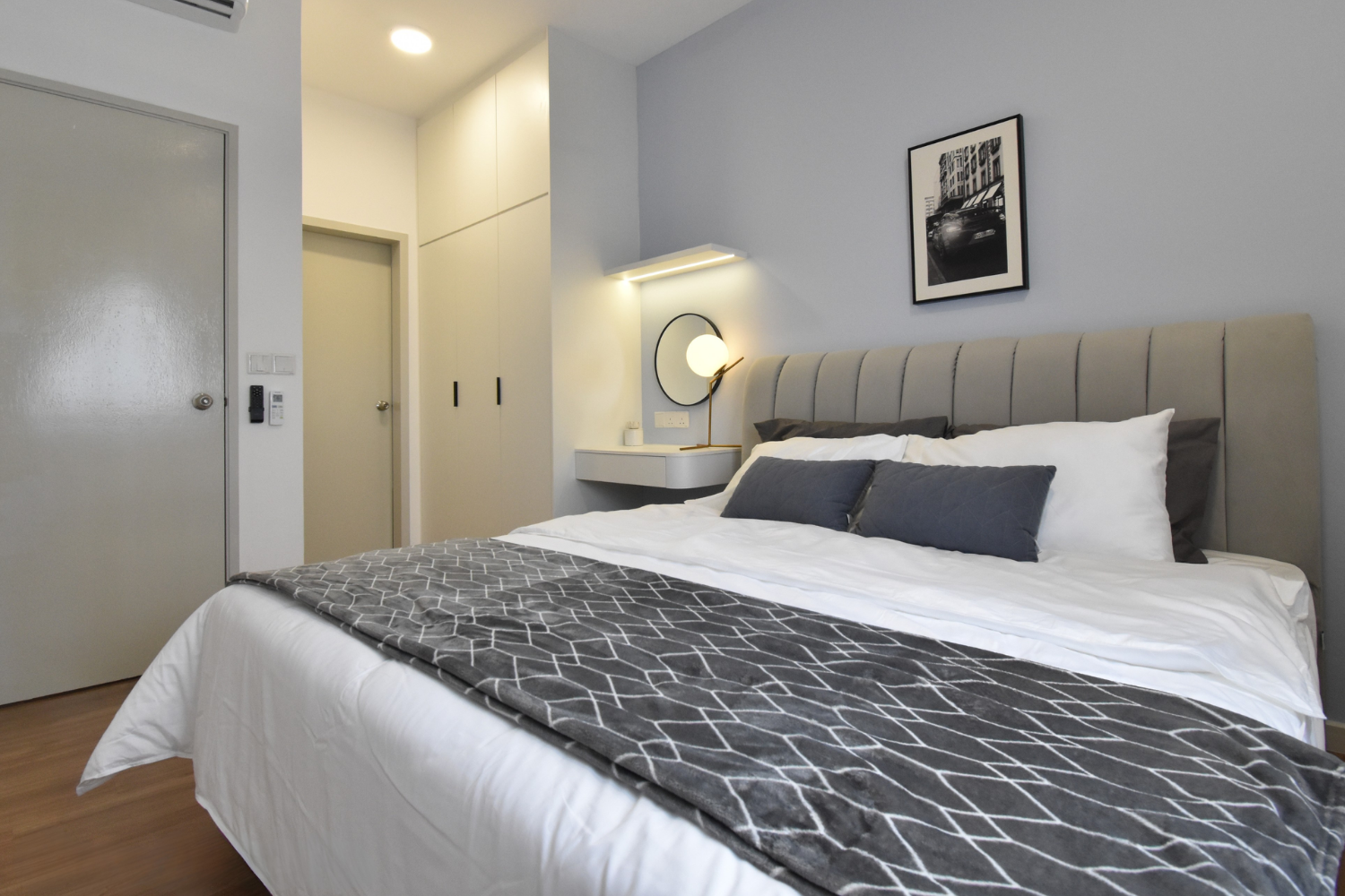 A neatly made bedroom with a large bed, gray headboard, white sheets, gray and black pillows, and a decorative gray blanket. The wall has a black and white cityscape photo, and there is a small white side table with a spherical lamp and a mirror on t