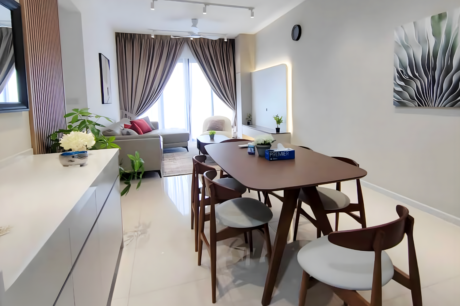 Modern living and dining room with a wooden dining table surrounded by six chairs, a white sideboard with decorative plants, a white sofa with cushions, a glass door with curtains behind the sofa, and decorative artwork on the walls.