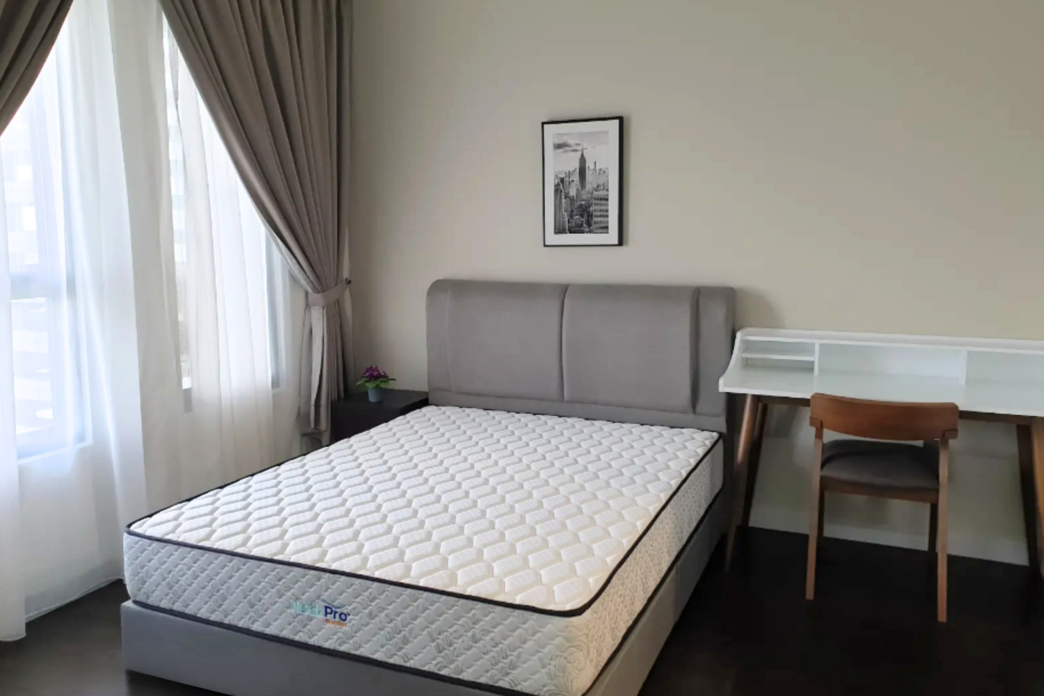A bedroom with a bed, grey upholstered headboard, nightstand with potted plant, framed black and white cityscape photo, white desk with shelves, wooden chair, and large window with white and beige curtains.