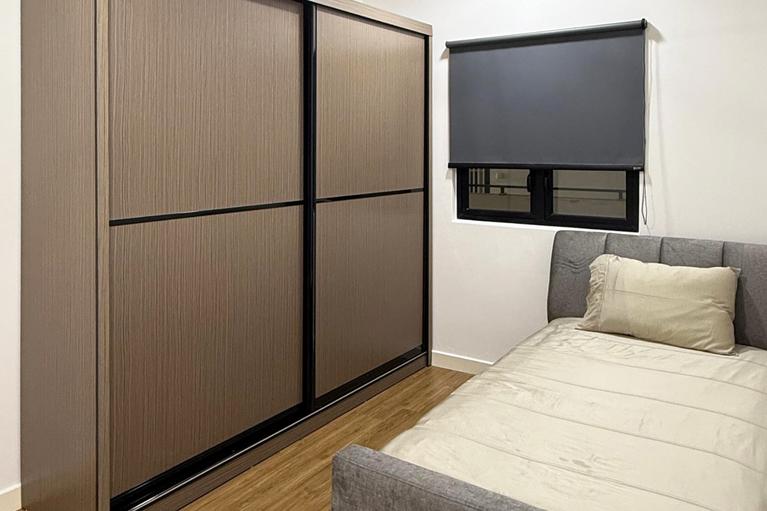 A bedroom corner featuring a grey upholstered bed with a beige pillow, a large closet with sliding doors framed in black, and a window with a closed dark grey roller shade. The room has light-colored walls and wooden flooring.