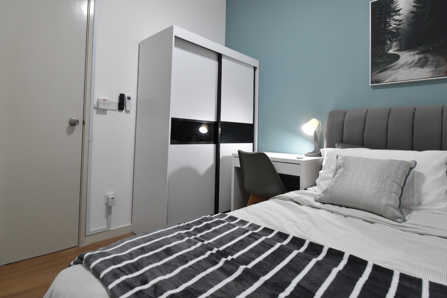 Bedroom with bed, grey headboard, striped blanket, pillow, bedside lamp, desk, grey chair, wardrobe, and wall art of a forest road.