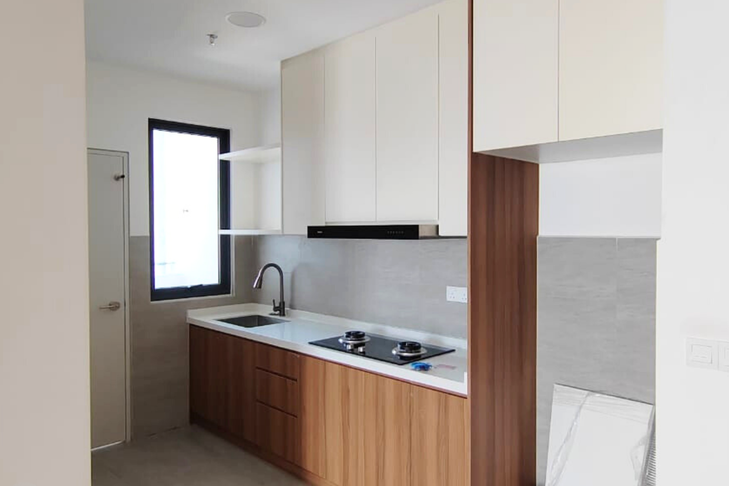 Modern kitchen with white cabinets, wooden lower cabinets, a black stovetop, and a black range hood. There is a sink with a black faucet, a window above the sink, and a door on the left. Light-colored tiles or backsplash behind the countertop.