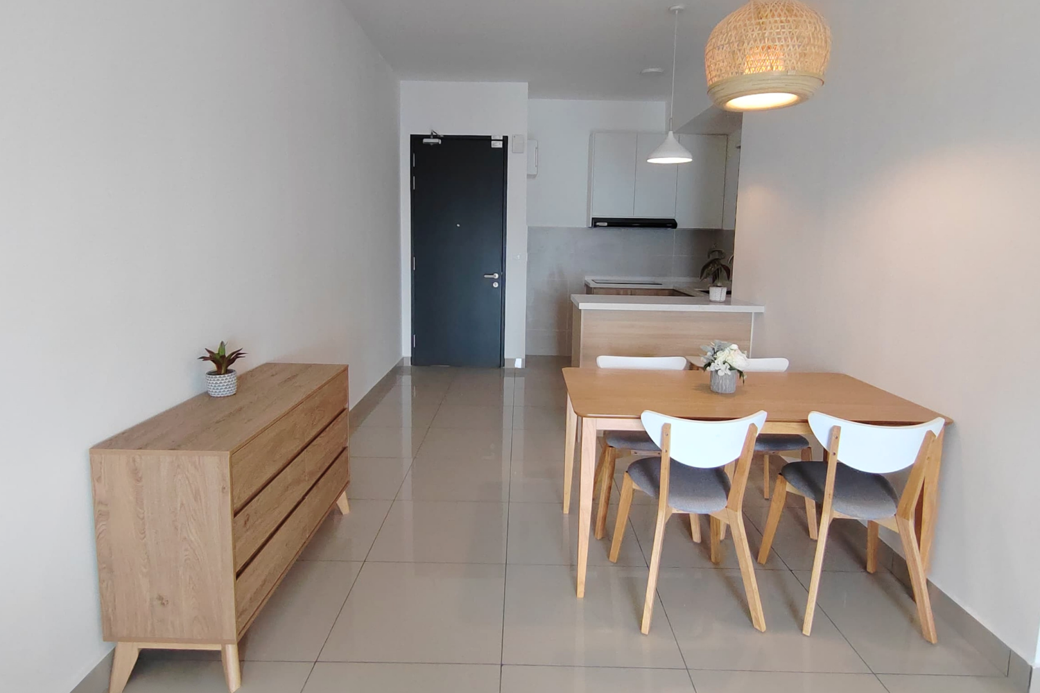 A dining area with a wooden table and four chairs, a wooden sideboard with a potted plant, and a kitchen in the background with white cabinets and a small potted plant on the counter. Two pendant lights hang above the table, and the space has light-c