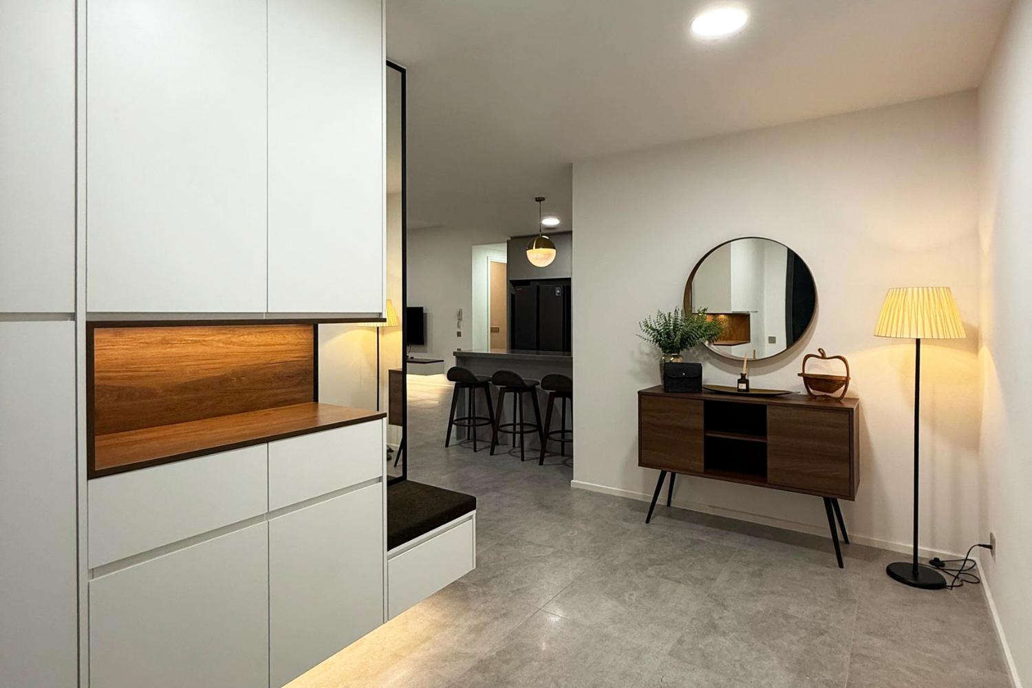 Modern living room with white and wood furniture, a round mirror on the wall, a potted plant, and a floor lamp emitting warm light, with a view of a kitchen and bar stools in the background.
