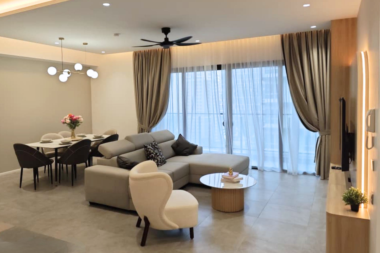 Living room with beige sofa, white armchair, round coffee table, dining table with flowers, large window with curtains, ceiling fan, and wall-mounted TV.