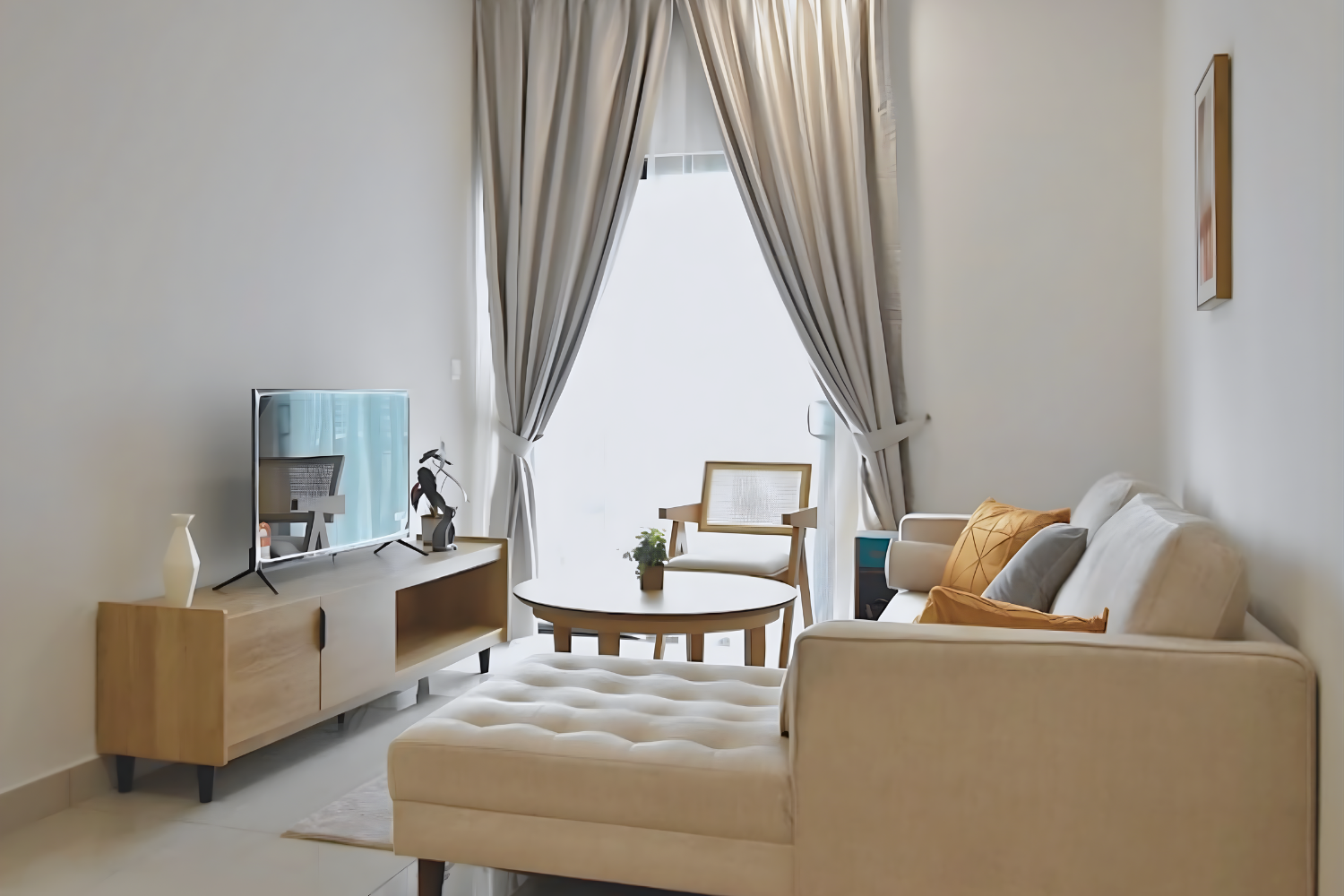Living room with a beige sofa, a round wooden coffee table, a TV on a wooden stand, a chair, and large window with beige curtains.
