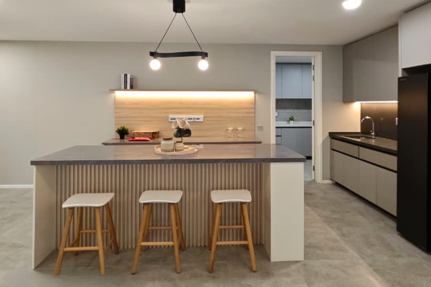 Modern kitchen with a central island featuring a gray countertop, three wooden barstools with white seats, a light wood-paneled back wall, a small plant, and decorative items, with an adjacent hallway and additional gray cabinetry and a black refrige