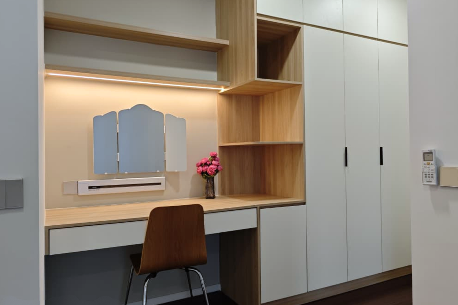 A modern built-in desk with a brown wooden chair and pink flower vase, beige wall with a small mirror, built-in wooden shelves, and a white cabinet unit.
