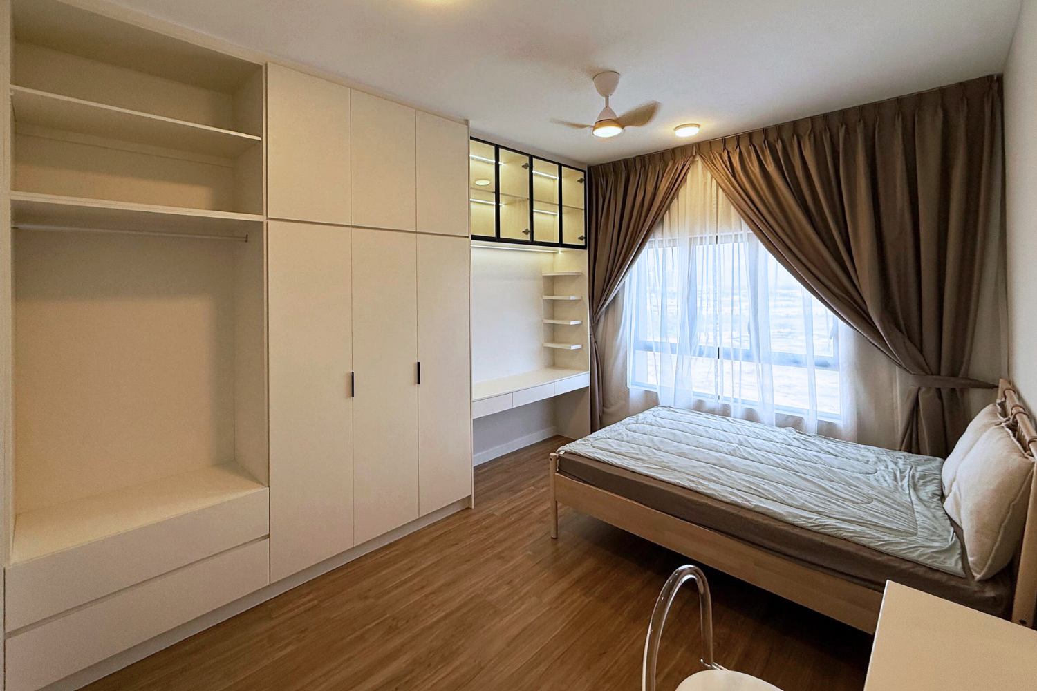 Empty bedroom with white built-in wardrobe, a desk, a bed with pillows, large window with brown curtains and sheer curtains, wooden floor, ceiling fan, and ceiling lights.