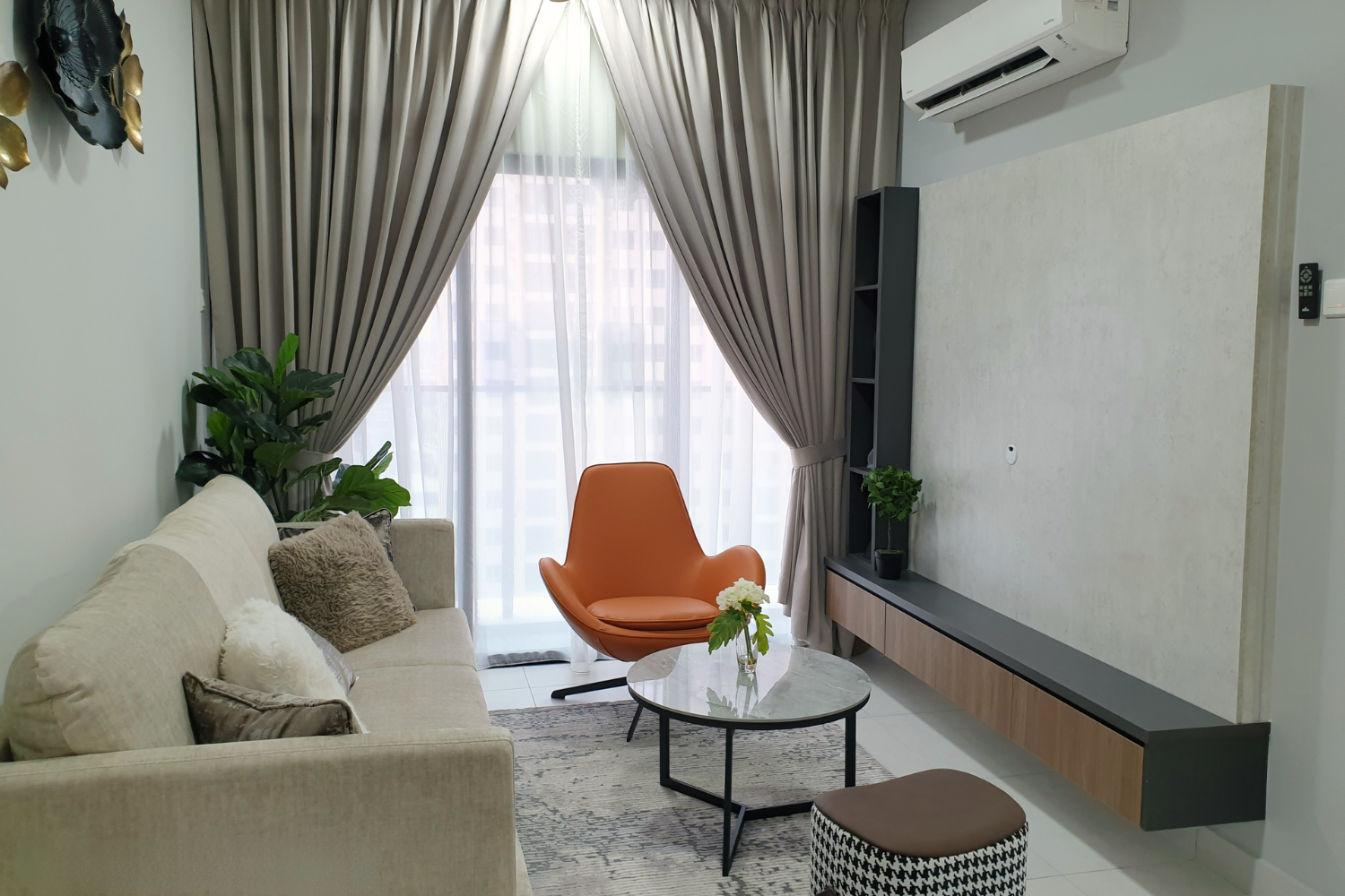 Modern living room with beige sofa, orange lounge chair, round coffee table with flower vase, large window with curtains, potted plants, and wall-mounted air conditioner.