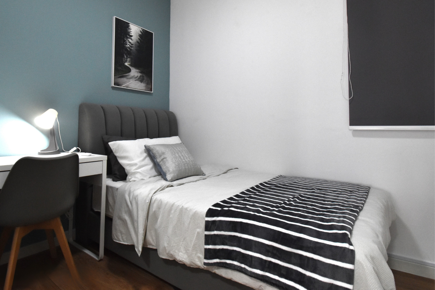 A modern bedroom with a bed, a desk with a lamp, and a framed black and white landscape photograph on the wall.