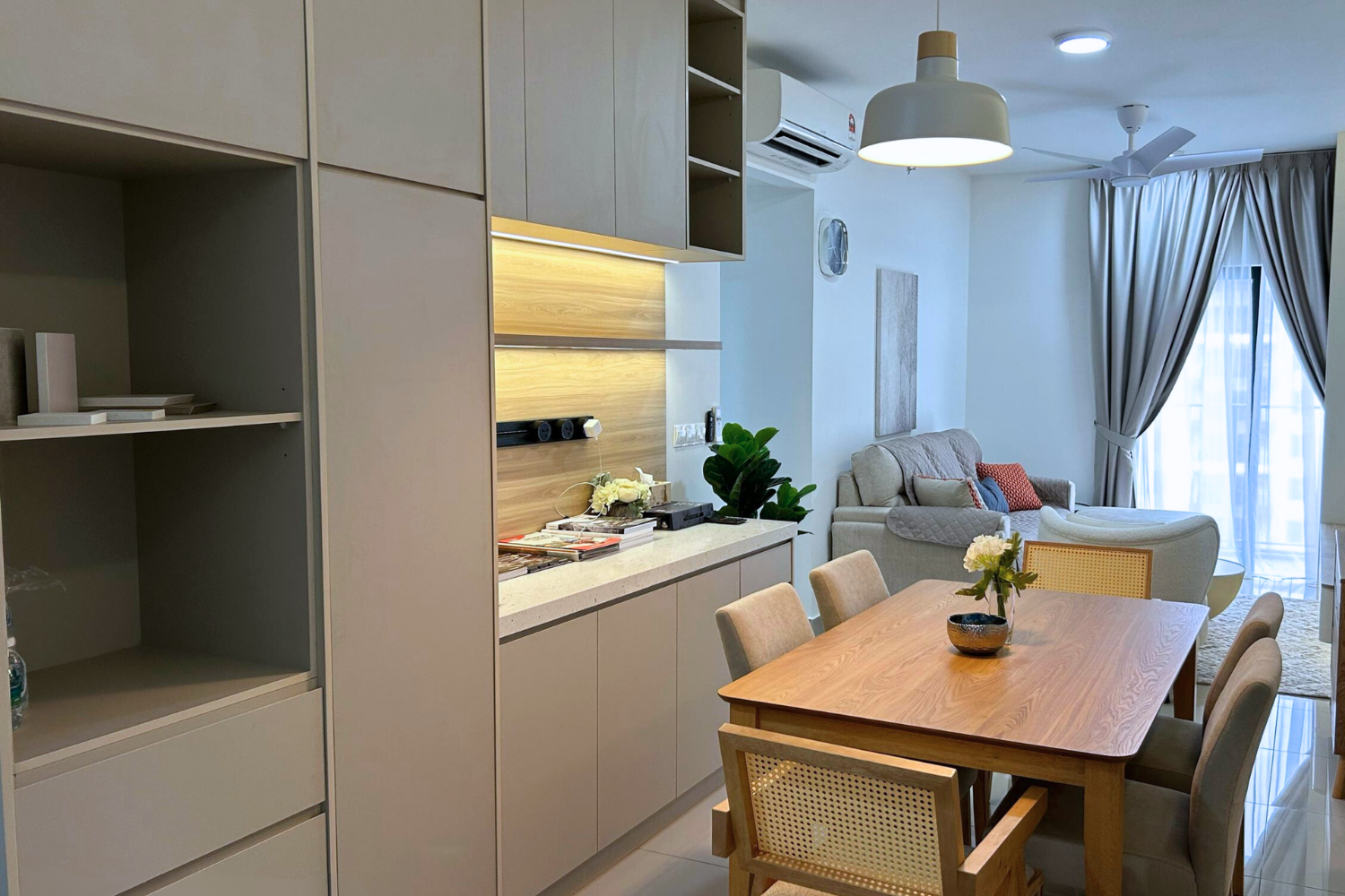 Living and dining area with beige sofa, wooden dining table with six beige chairs, vase with white flowers, grey curtains, blue cushion, cream-colored walls, ceiling fan, air conditioner, pendant light, and balcony with glass door.