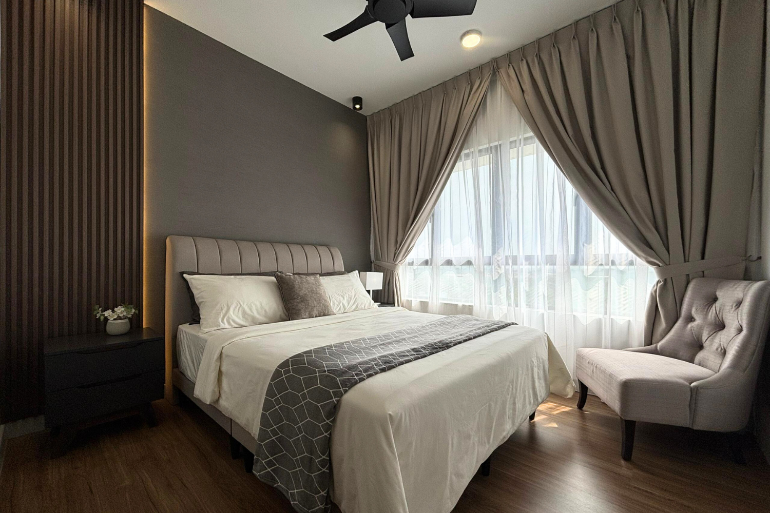 Modern bedroom with a bed, gray headboard, beige curtains, sheer curtains, a tufted armchair, and a nightstand with a flower vase.