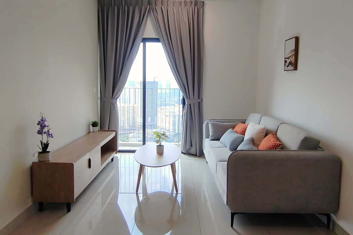 Modern living room with a gray sofa with decorative pillows, a small round coffee table with a potted plant, a long wooden TV stand with a potted orchid and plant, large glass sliding doors with gray curtains, overlooking a city skyline.