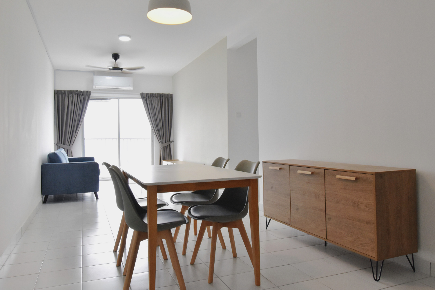 Modern living and dining area with a wooden dining table, black chairs, a wooden sideboard, gray curtains, a blue sofa, and a balcony with a sliding glass door.