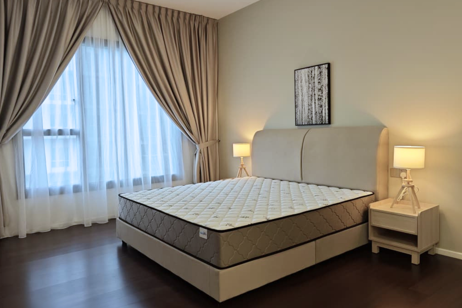 A bedroom with a large bed, a beige headboard, two nightstands with lamps, beige curtains, and a window with sheer white curtains.