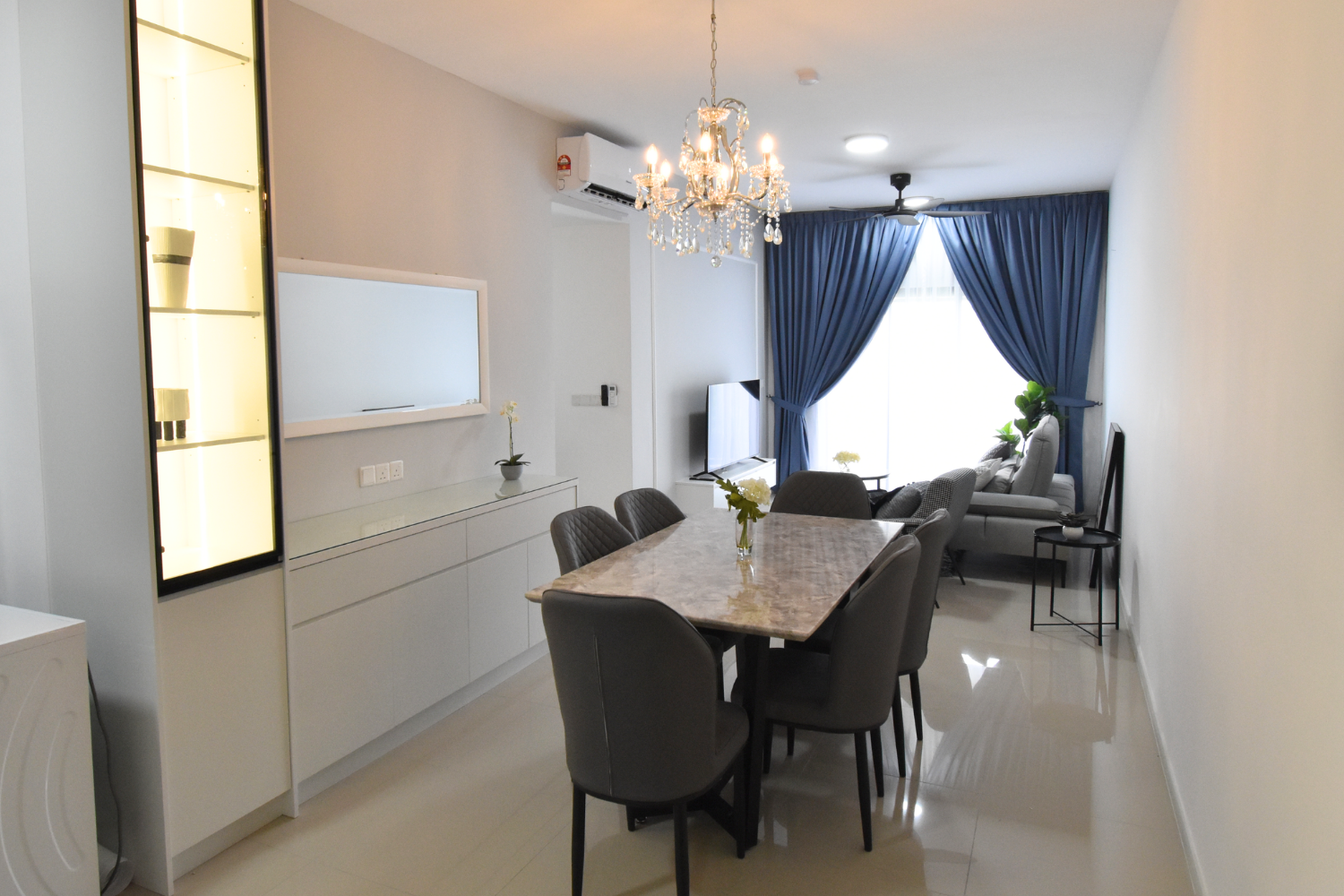 Living and dining room with white walls, blue curtains, chandelier, gray and beige furniture, and a television.