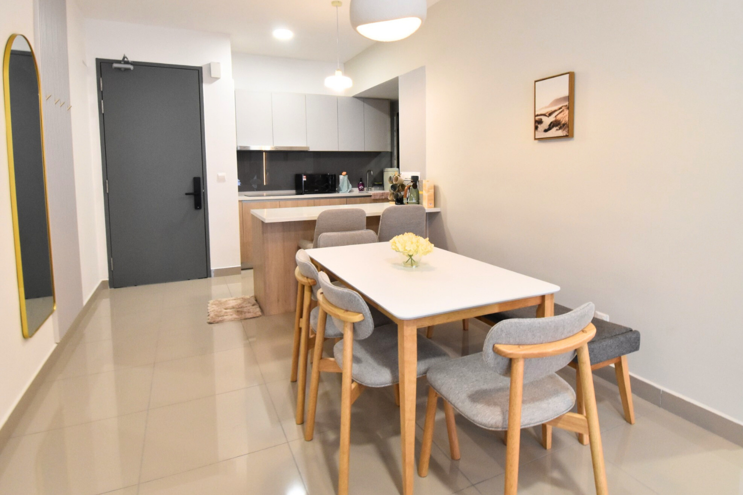 Dining area with a white table, six wooden and grey upholstered chairs, a vase of yellow flowers, and a view of the kitchen in the background with white cabinets, a black countertop, and various appliances.