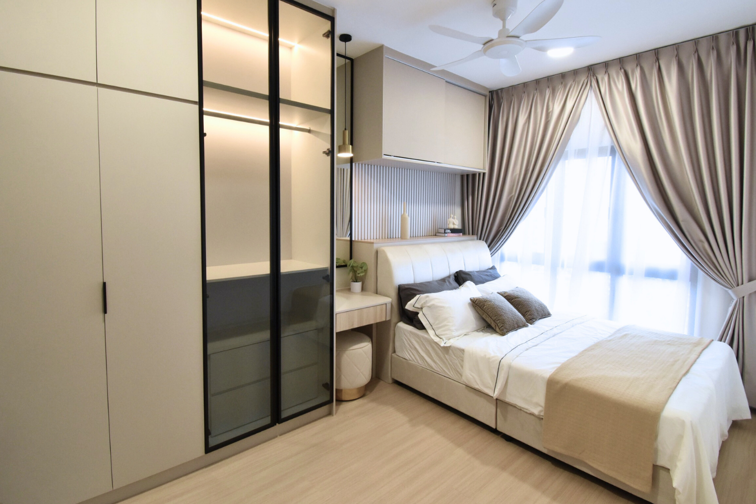 A modern bedroom with a bed, decorative pillows, curtains, a ceiling fan, a large wardrobe with glass doors, and a small vanity table with a potted plant and decorative items.