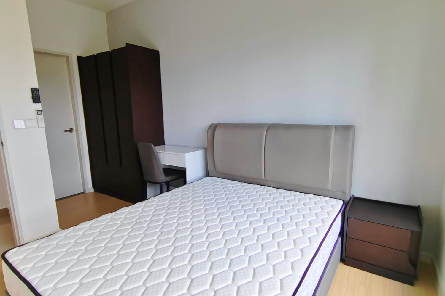 Empty bedroom with a large bed, beige headboard, small black nightstand, dark wardrobe, desk with a gray chair, plain white wall, and light wood floor.