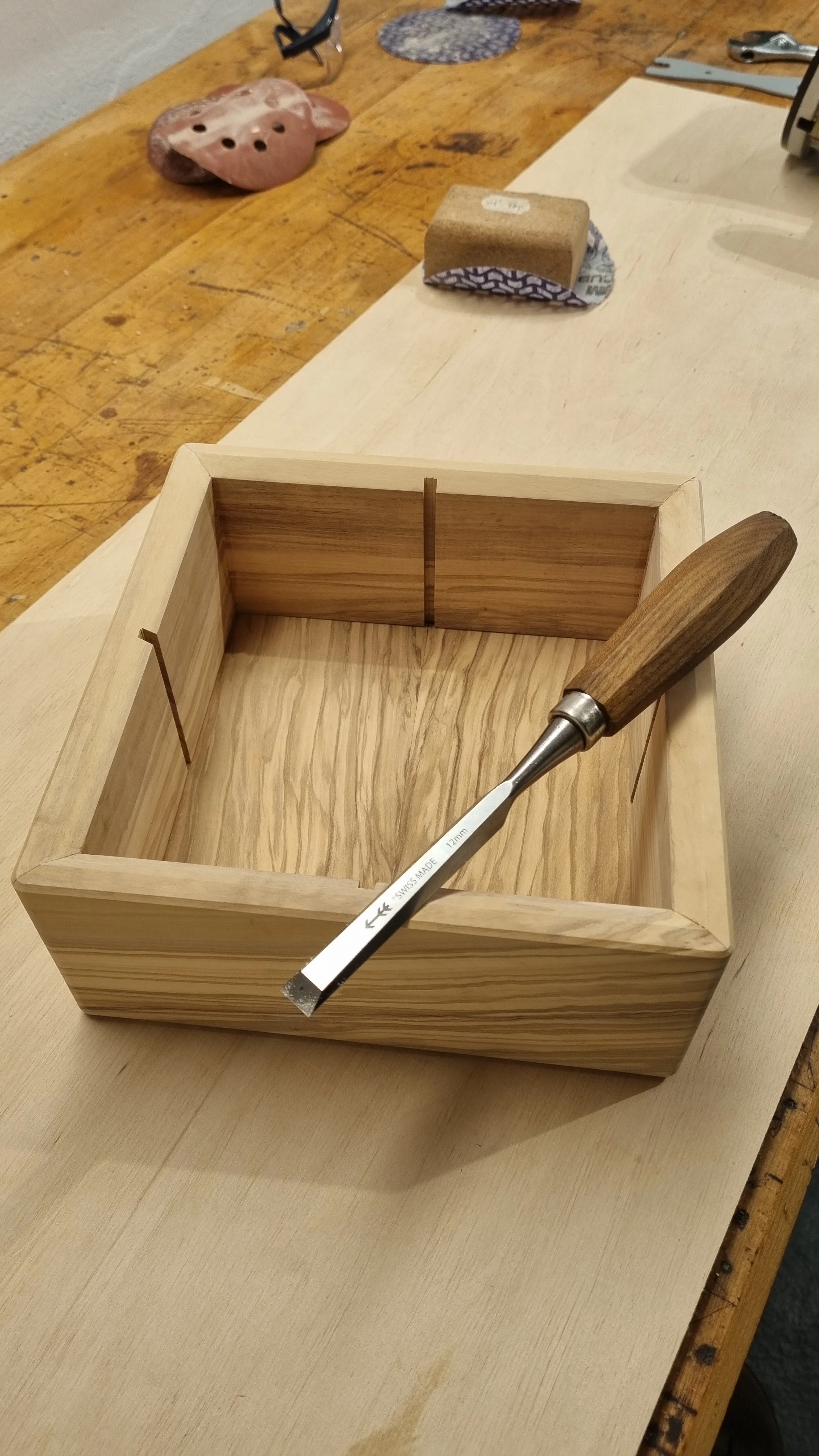 Ein leerer Holzkasten auf einem Tisch, mit einem Meißel darin, in einem Holzwerkstatt-Umfeld.