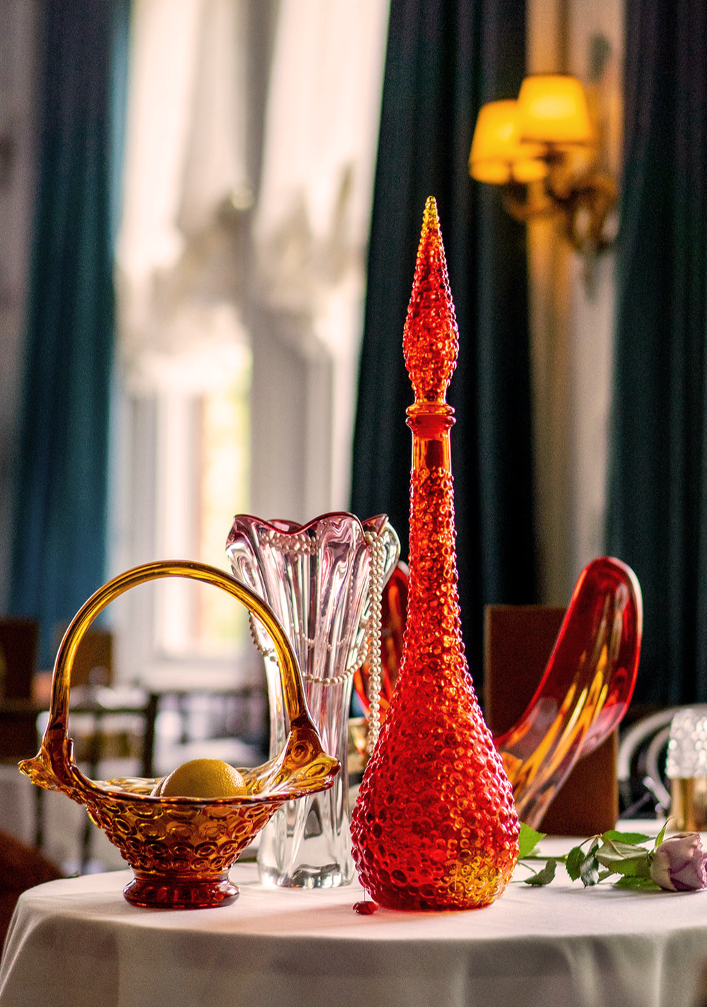 Decorative glass vases and bowl on a table with a white tablecloth, with a flower and drapes in the background.